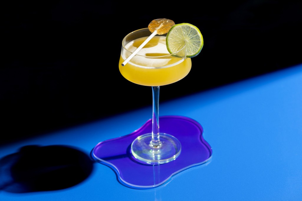 A Coral Crush cocktail garnished with a slice of lime on a purple table