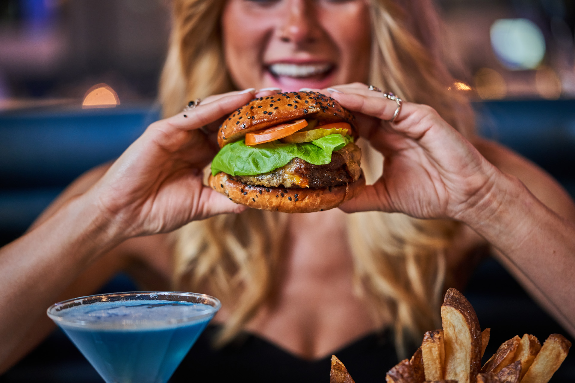 A blonde woman with a big smile on her face looking at a big succulent burger which she is holding with both hands.