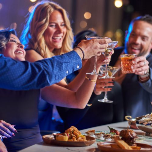 A group of friends are cheersing cocktails together. One man is holding an old fashioned, whilst the three women have martinis