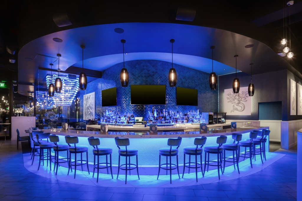 Interior shot of a bar with curved counter illuminated by blue lighting, high backed bar stools, multiple pendant lights, and a textured blue tile wall with three large blank screens behind the bar.